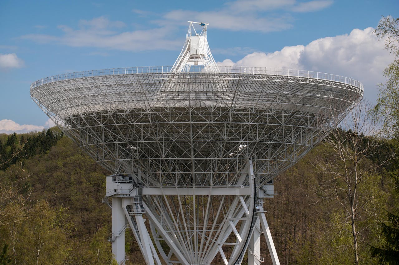 creative-03 A large radio telescope set against a backdrop of trees and clouds, capturing celestial signals.